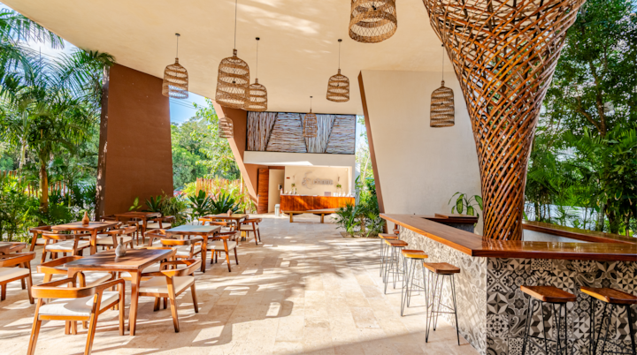 Une terrasse d'hôtel élégante avec des meubles modernes en bois, des suspensions en osier et un bar orné de carreaux aux motifs travaillés, le tout au milieu de plantes d'un vert éclatant.