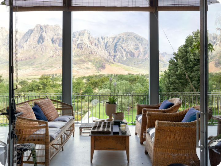 Une terrasse paisible avec des meubles en osier, une table basse et des plantes en pot, surplombant un jardin luxuriant et des montagnes lointaines sous un ciel nuageux.