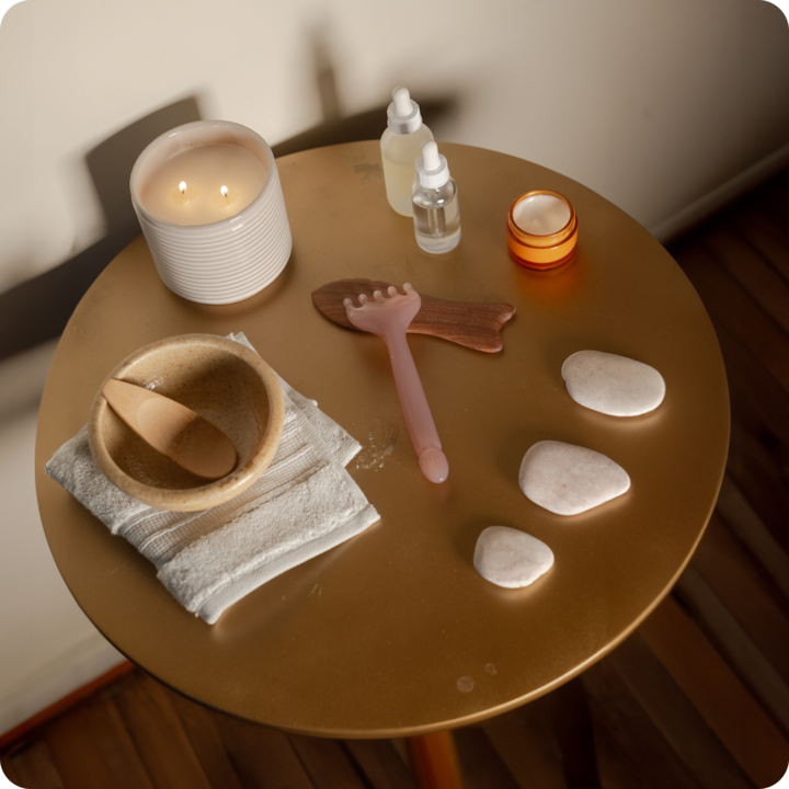 Une ambiance de spa relaxante avec une bougie allumée, des flacons, un bol en bois, une serviette, un outil de massage rose et des pierres blanches posés sur une table.