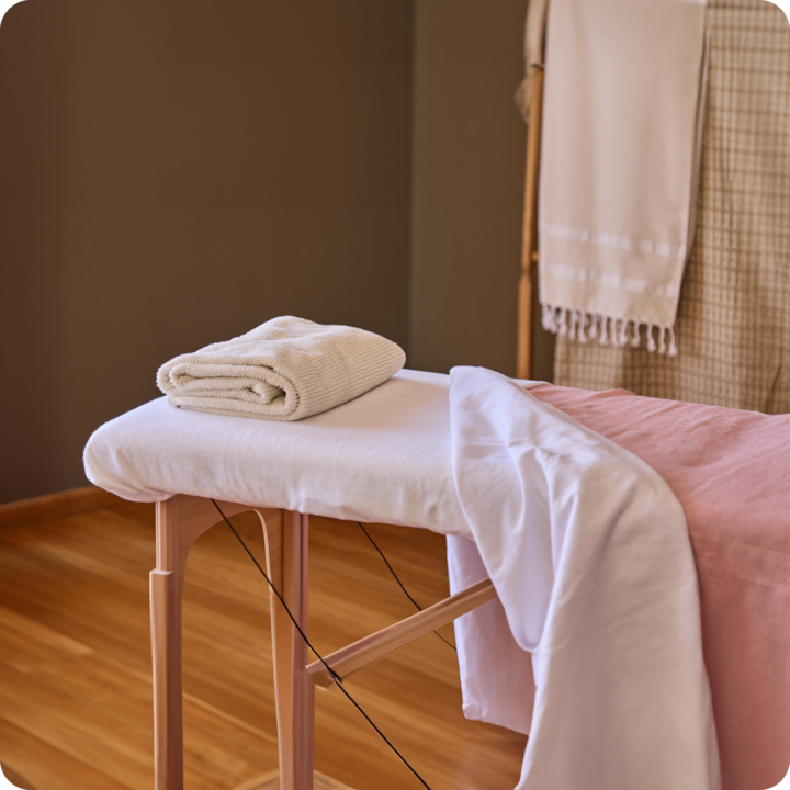 Un espace bien-être paisible, équipé d'une table de massage protégée par un drap sur laquelle est posée une serviette pliée, le tout dans une pièce aux tons doux et neutres.