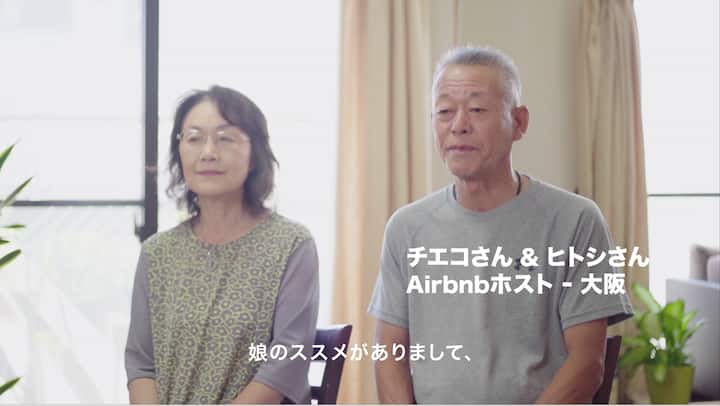 Two people are seated indoors in a bright room with large windows and beige curtains, with Japanese text overlay describing them as Airbnb hosts in Osaka.