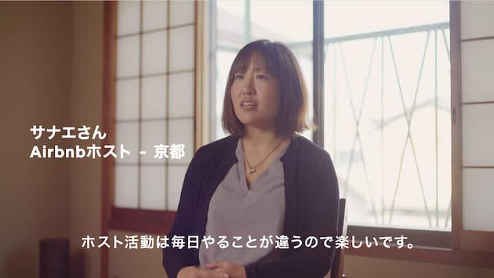 A woman seated in a traditional Japanese room with shoji screens and a window in the background. Text on the image reads: 'サナエさん Airbnbホスト - 京都' and 'ホスト活動は毎日やることが違うので楽しいです.'