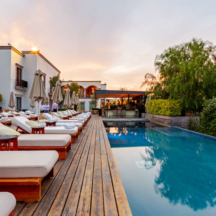 Un espace extérieur élégant d'un hôtel, avec des chaises longues au bord de la piscine, une terrasse en bois et une piscine à débordement paisible qui reflète un ciel coloré au coucher du soleil.