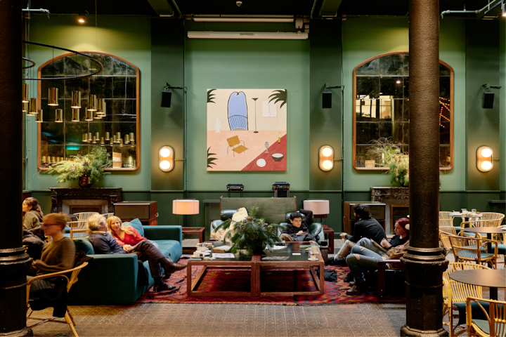 Un salon d'hôtel élégant aux murs verts, avec de grands miroirs et un décor moderne. Des clients se détendent sur des fauteuils et canapés confortables, entourés de plantes vertes et baignés d'une lumière douce.
