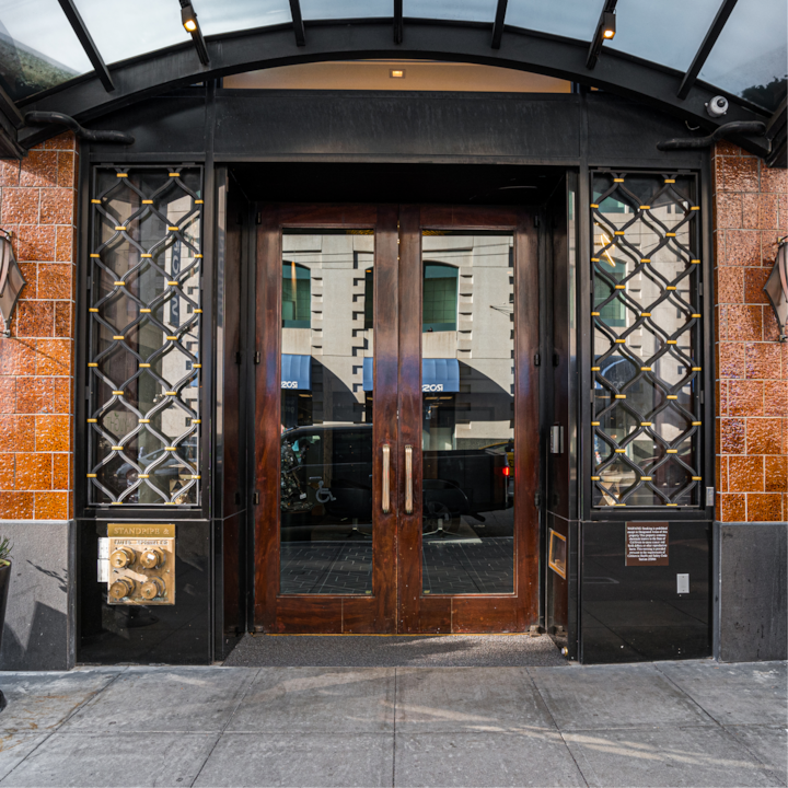 Une entrée d'hôtel élégante avec de grandes portes doubles en bois, des treillis métalliques complexes des deux côtés et un auvent en verre au-dessus, reflétant une scène de rue en ville.