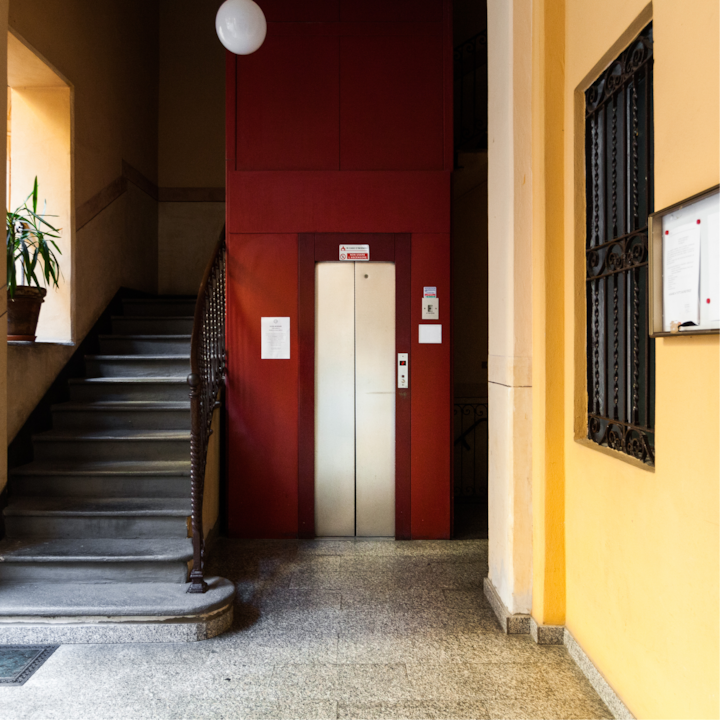 Un hall d'immeuble chaleureux avec une porte d'ascenseur rouge au centre, un escalier en colimaçon avec une balustrade en bois sur la gauche et des murs jaunes qui créent une atmosphère accueillante.
