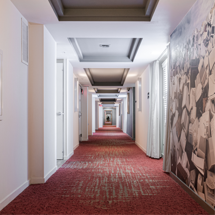 Un long couloir d'hôtel avec une moquette rouge à motifs, des murs blancs et des moulures grises au plafond, menant à une porte distante.