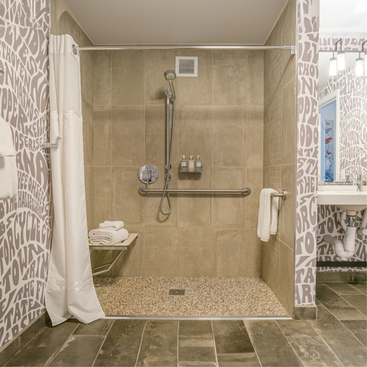 Douche à l'italienne accessible au carrelage beige, avec des barres d'appui, une douchette à main, des articles de toilette fixés au mur et un banc pliable sur lequel des serviettes sont soigneusement empilées.