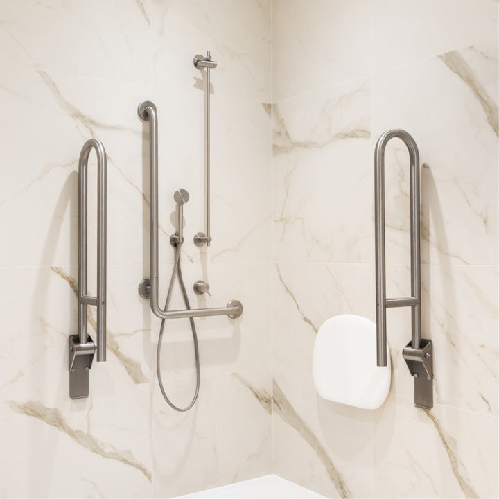Une douche lumineuse et accessible avec des murs en marbre poli, dotée d'un pommeau de douche fixé au mur, de barres d'appui et d'un siège de douche pliable.