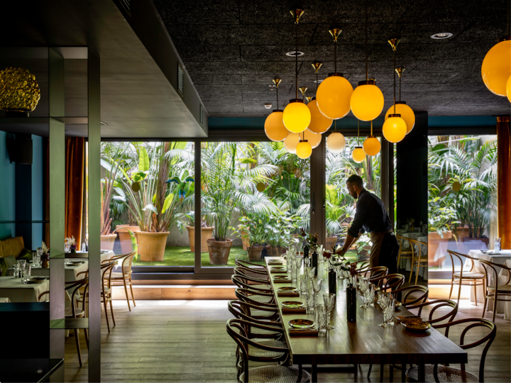 Un espace repas baigné d'une lumière chaleureuse, avec une longue table en bois dressée et entourée de chaises élégantes, ainsi que de grandes baies vitrées donnant sur des plantes tropicales luxuriantes.