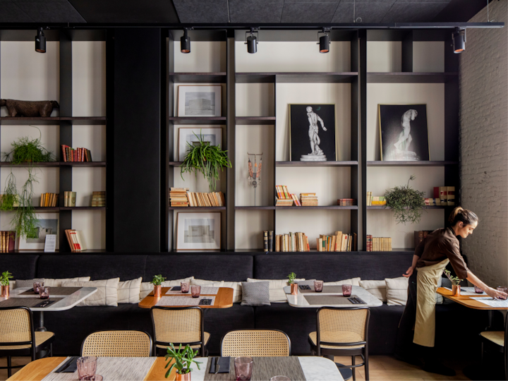 Un espace restaurant élégant avec des tables en bois, des chaises en rotin et une grande étagère remplie de livres, de plantes et d'œuvres d'art. Une personne en tablier ajuste le dressage d'une table.