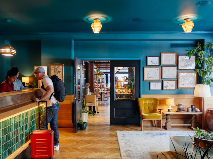 Un hall d'hôtel accueillant avec des murs d'un bleu canard éclatant, un parquet en bois et une lumière chaleureuse. Un homme avec un sac à dos et une valise rouge s'enregistre à la réception, un espace accueillant décoré de cadres et de plantes. La porte ouverte donne sur un espace repas bien éclairé, équipé de tables et de chaises.