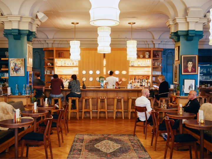 Un bar d'hôtel accueillant avec des colonnes ornées, une décoration colorée et des sièges confortables. Des clients assis à des tables ou au bar se détendent, sous des suspensions à la lumière douce qui créent une atmosphère accueillante.