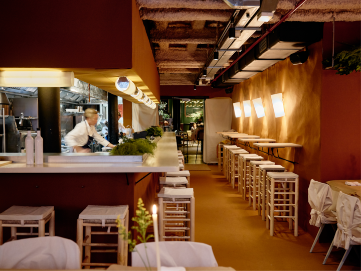 Intérieur d'un restaurant intimiste aux tons ocres, avec des appliques murales lumineuses et un chef qui prépare des plats derrière un comptoir. La salle à manger est aménagée avec des tabourets en bois et des tables bien disposées.