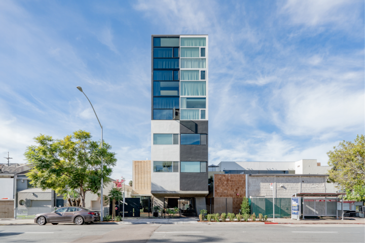 Un hôtel contemporain à l'architecture géométrique, doté de hautes fenêtres et d'un design épuré, niché dans un cadre urbain avec des arbres et des voitures en stationnement à proximité.
