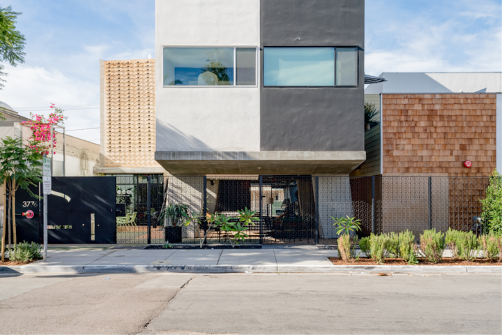 Extérieur d'un boutique-hôtel moderne au design géométrique, avec des textures contrastées, notamment du béton gris lisse, du bois tressé et du verre. Une végétation luxuriante borde l'entrée.