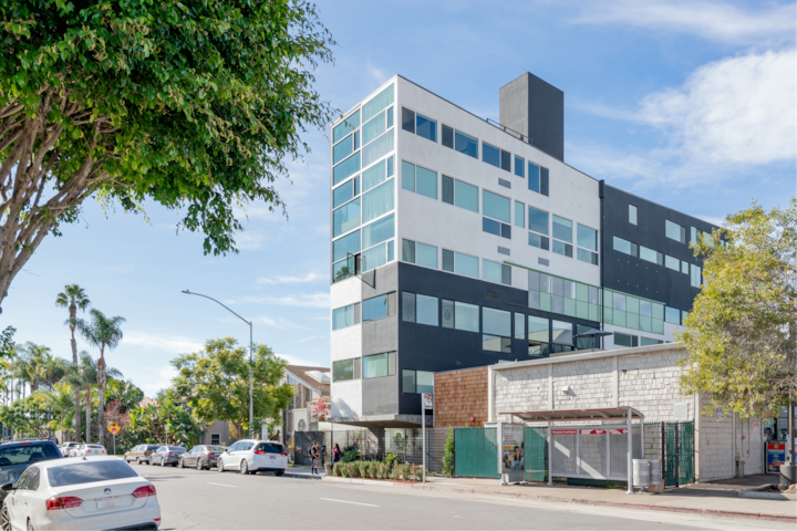 Vue extérieure d'un hôtel élégant avec une façade contrastée en noir et blanc, de grandes fenêtres et un environnement urbain : arbres, voitures en stationnement et arrêt de bus.