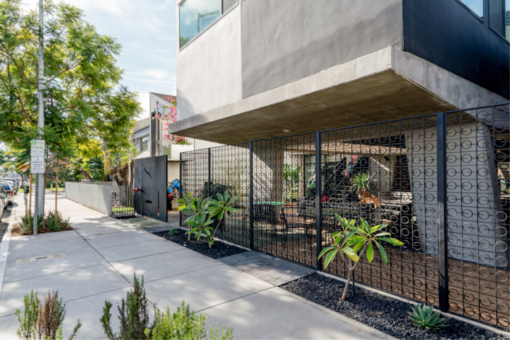 Extérieur lumineux d'un bâtiment élégant, encadré par une clôture géométrique, un aménagement paysager tropical et un trottoir bien entretenu dans un quartier animé.