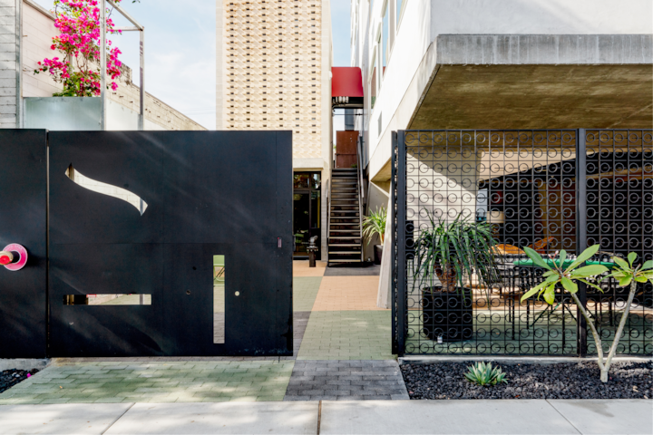 Une façade d'hôtel élégante avec un portail noir épuré, des fleurs roses en hauteur, des murs en briques à motifs et un salon extérieur accueillant entouré de plantes.