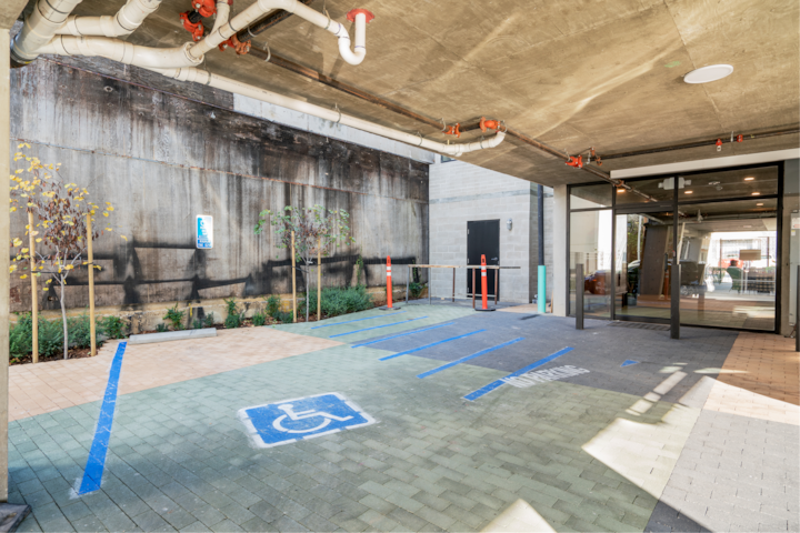 Parking couvert avec des places de stationnement accessibles identifiées en bleu, et entourées de petites plantes et d'arbres, près de l'entrée d'un bâtiment.
