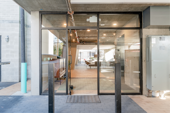 Entrée d'un bâtiment contemporain avec des portes vitrées, révélant un intérieur de style industriel doté de sièges de salon et d'un éclairage chaleureux.