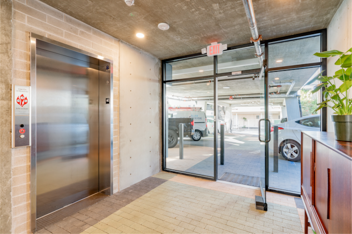 Hall lumineux de style industriel avec un ascenseur et des portes vitrées donnant sur un parking extérieur où l'on aperçoit des véhicules.