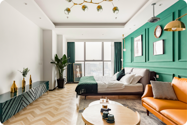 Une chambre d'hôtel lumineuse et moderne, avec un mur peint en vert orné de tableaux, un lit confortable près d'une grande fenêtre, un élégant canapé orange et un parquet en chevron.