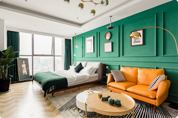 Une chambre d'hôtel contemporaine aux couleurs vives, avec un mur vert à moulures orné de cadres, un lit soigneusement fait et un canapé orange accompagné d'une table basse élégante. Une grande fenêtre laisse entrer la lumière du jour et la ville se dessine en arrière-plan.