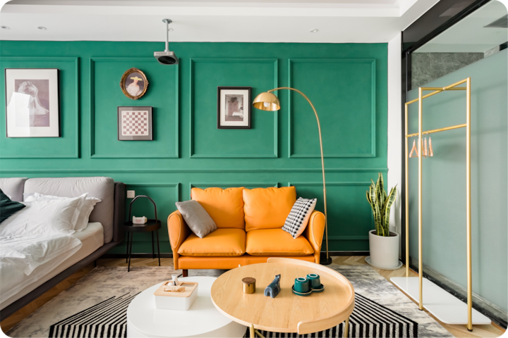Un espace accueillant avec un mur peint en vert vif orné d'œuvres d'art, un canapé jaune confortable, une table basse ronde, un tapis à rayures et un portant à vêtements minimaliste à côté d'une plante en pot.
