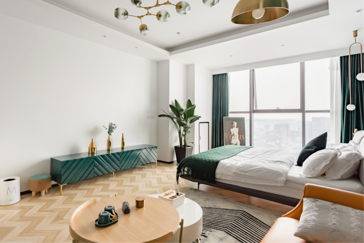Une chambre d'hôtel moderne au design minimaliste, baignée de lumière naturelle, avec un lit luxueux, des éléments de décoration verts et dorés, une plante en pot et une grande fenêtre donnant sur la ville.