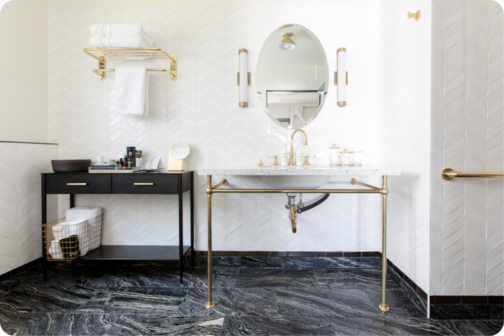 Une salle de bain blanche élégante, dotée d'un lavabo en marbre à la robinetterie dorée, d'un mur carrelé à chevrons et d'une console noire sur laquelle sont soigneusement disposés des serviettes et des produits de toilette.