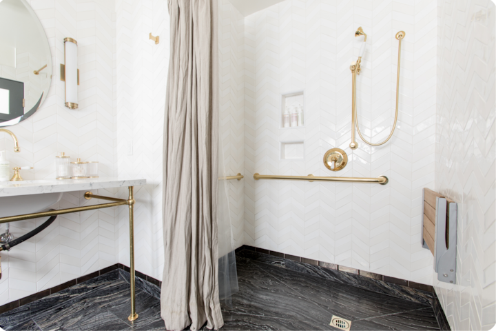 Une salle de bain lumineuse agrémentée de touches dorées, avec lavabo en marbre, sol effet ardoise et équipements d'accessibilité, notamment une douche à l'italienne avec barre d'appui.