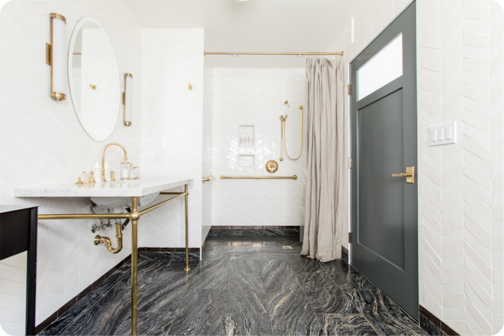 Une salle de bain élégante aux accents dorés, dotée d'un lavabo en marbre, d'un miroir ovale, d'une douche à l'italienne avec des barres d'appui et d'un sol noir texturé.