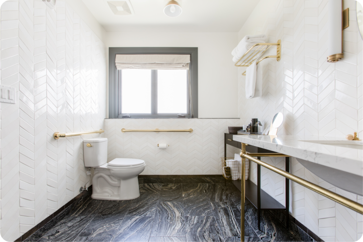 Une salle de bain accessible comprenant des toilettes et un lavabo spacieux dotés de barres d'appui dorées, et des murs en carrelage blanc à chevrons.