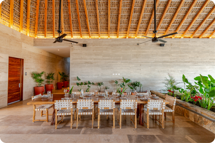 Un coin repas spacieux avec une longue table en bois entourée de chaises en tissu, dressée pour un repas. La pièce dispose d'un toit de chaume et de murs en pierre claire. Elle est agrémentée de jardinières contenant une végétation luxuriante sur le côté, ce qui crée une ambiance tropicale.