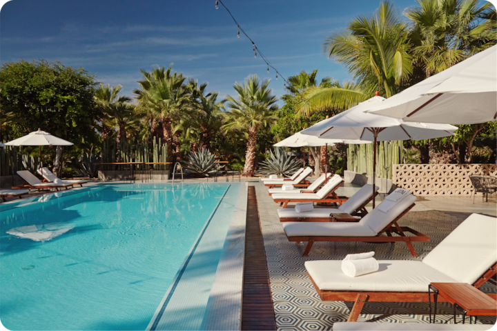 Piscine extérieure entourée de palmiers et d'une végétation luxuriante, avec des chaises longues et des parasols sur une terrasse en carrelage à motifs, sous un ciel bleu dégagé.