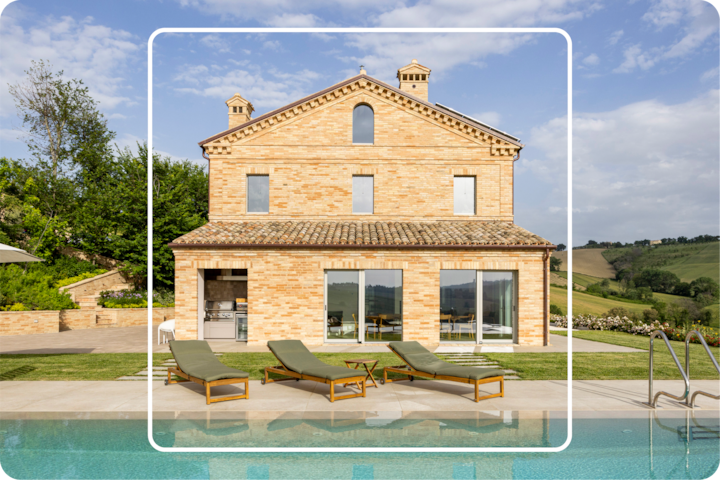 Une villa élégante en briques au design classique, avec piscine extérieure et chaises longues, dans un cadre avec verdure et champs sous un ciel en partie nuageux.