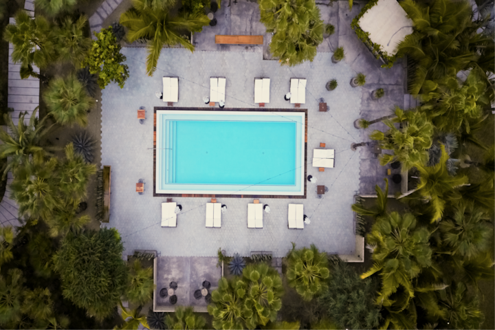Vue aérienne de l'espace piscine d'un complexe hôtelier, avec une piscine à l'eau turquoise au centre, des chaises longues soigneusement disposées et une végétation luxuriante.