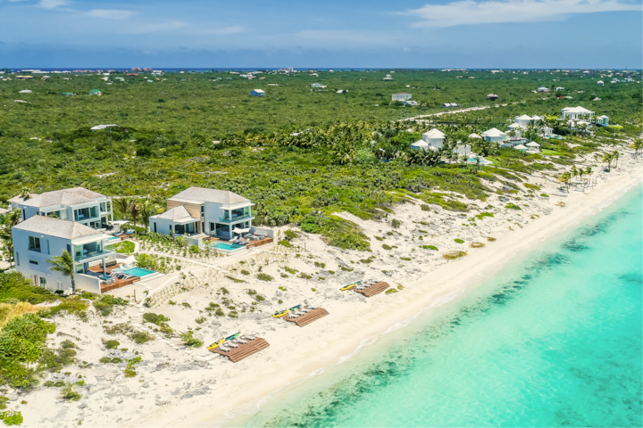 Vue prise par drone de villas en bord de mer dotées de piscines privées, nichées le long d'un rivage de sable blanc aux eaux turquoise, entourées d'une végétation luxuriante.