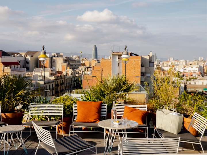 Un toit-terrasse avec des chaises blanches modernes, des coussins orange et des plantes en pot, surplombant un paysage urbain mêlant bâtiments en briques ou contemporains sous un ciel bleu.