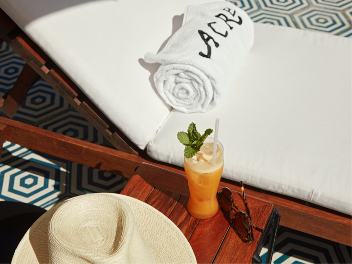 Un moment de détente au bord de la piscine mis en scène avec une chaise longue blanche, une serviette portant la marque « Acre », un chapeau de paille, des lunettes de soleil et une boisson à l'orange agrémentée de menthe posée sur une petite table en bois.