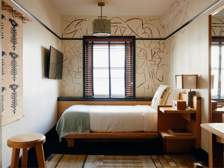 Une chambre confortable et bien éclairée, dotée d'un lit simple, de murs à motifs abstraits, d'une grande fenêtre avec des stores et d'un mobilier minimaliste en bois.