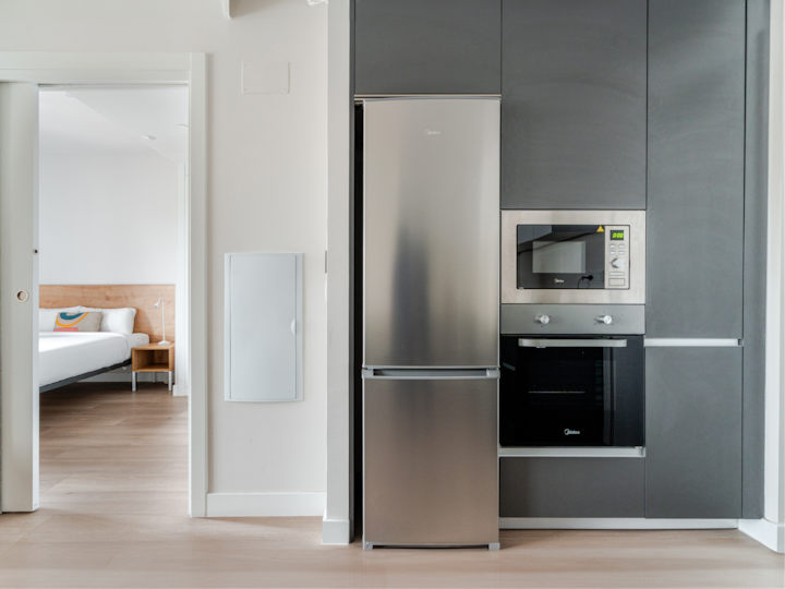 Cuisine contemporaine équipée de placards sombres et d'appareils électroménagers en acier inoxydable, avec une porte donnant sur une chambre lumineuse à la décoration épurée et moderne.