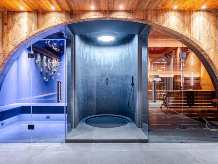 Espace spa luxueux comprenant une douche à effet de pluie circulaire avec des murs gris épurés, un bassin de relaxation éclairé en bleu à gauche et un sauna finlandais aux tons chauds de bois à droite, créant ainsi une atmosphère sereine et élégante.
