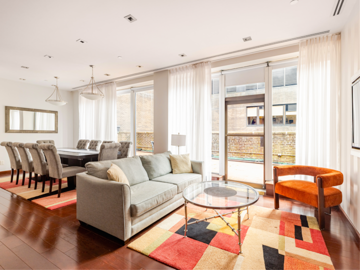 Une pièce lumineuse et élégante avec de grandes baies vitrées, dotée d'un coin salon confortable composé d'un canapé gris et d'un fauteuil orange, ainsi que d'une table à manger pour huit personnes avec des chaises confortables, le tout reposant sur un tapis à motifs.