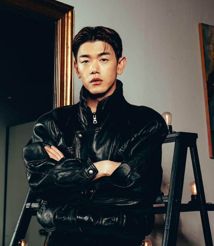 A person in a stylish black leather jacket stands in a studio setting, with vintage decor including a framed mirror and ladder adorned with light bulbs, evoking an artistic and moody atmosphere.