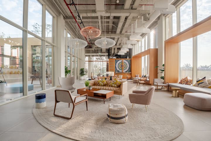 Un salon spacieux et moderne avec de grandes fenêtres, du mobilier aux tons neutres, des plafonniers artistiques et une fresque colorée sur le mur du fond.