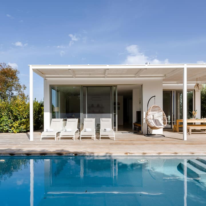 Maison contemporaine dotée de grandes portes coulissantes en verre donnant sur une terrasse avec des chaises longues, un fauteuil suspendu et une piscine, sous un ciel bleu vif.