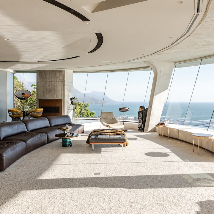 L'intérieur d'un espace de vie lumineux et ouvert avec des baies vitrées incurvées offrant une vue panoramique sur l'océan, doté de sièges élégants et d'éléments de décoration minimalistes.