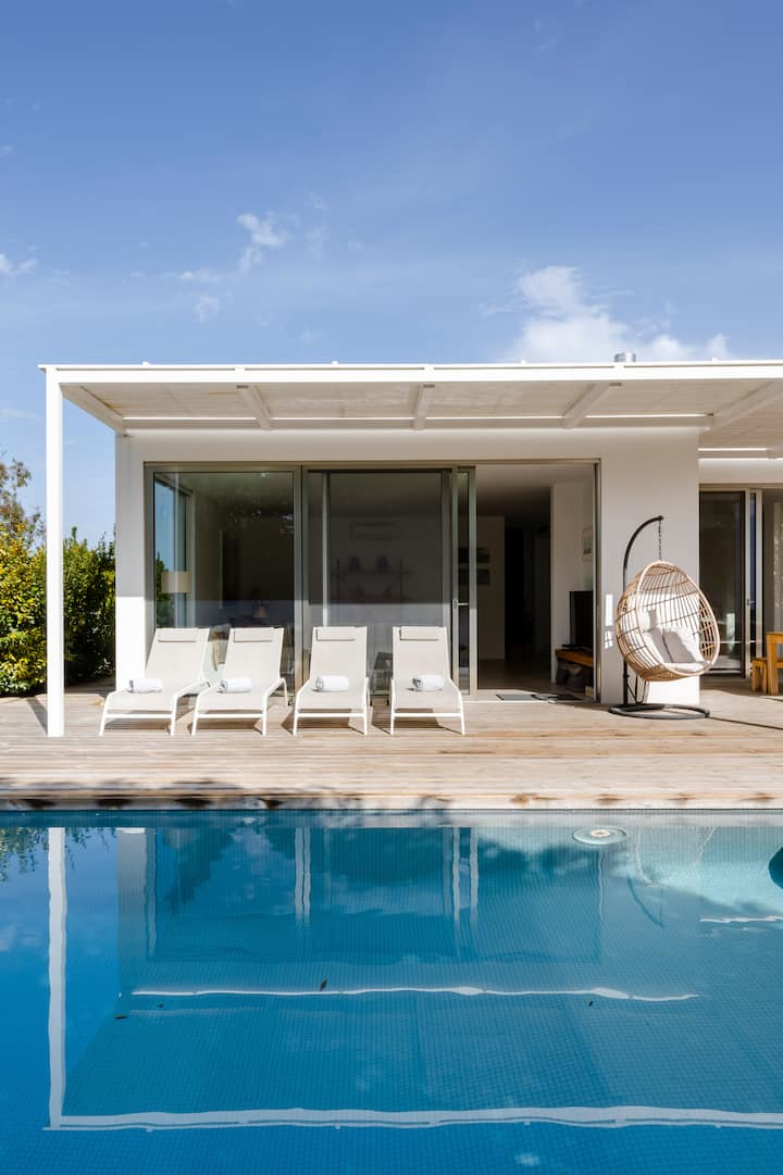 Un espace au bord d'une piscine avec des chaises longues blanches, des serviettes roulées et une chaise en osier suspendue. Une maison blanche moderne, dotée de portes coulissantes en verre et d'une terrasse spacieuse, se trouve à l'arrière-plan.